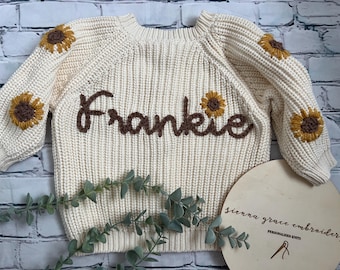 May include: A cream-colored knitted sweater with brown embroidered sunflowers and the name "Frankie" on the front.