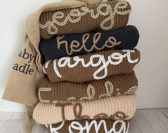 Può includere: Una pila di coperte per bambini personalizzate in varie tonalità di marrone e beige, ciascuna ricamata con un nome in filo bianco. Una testa di coniglio di peluche e una mussola con la scritta "baby cradle" sono in cima.