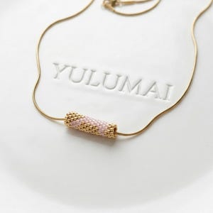 Può includere: Una delicata collana dorata con un pendente a forma di cilindro composto da perline rosa e dorate. La collana è appoggiata su una superficie bianca con la scritta "YULUMAI" in rilievo. Il design del pendente è ripetitivo.