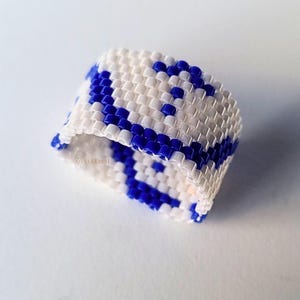 Peut inclure: Une bague faite à la main en perles avec un motif répétitif de perles bleues et blanches. La bague est de style large, avec un design géométrique. Les perles sont petites et étroitement tissées, créant une surface texturée. La bague est probablement un accessoire de mode.