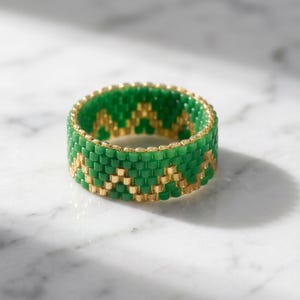 May include: A beaded ring constructed from small cylindrical green and metallic gold beads. The beads are arranged in a repeating zigzag or chevron pattern that encircles the entire band, creating a textured surface with a consistent width.