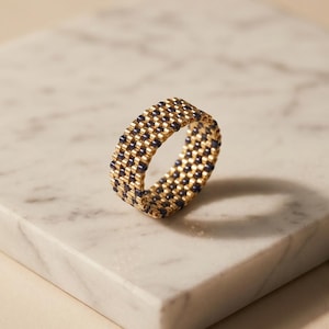 Peut inclure: Une bague tissée présentant un motif répétitif de perles dorées métalliques et bleu marine foncé. L'anneau est composé de petites perles cylindriques uniformes disposées en grille, créant un aspect texturé et flexible.