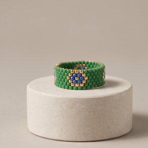 Peut inclure: Une bague en perles composée de petites perles cylindriques vertes disposées en anneau. Le motif présente une répétition d'une perle bleue centrale entourée d'un cercle de six perles dorées métalliques sur un fond de perles vertes.