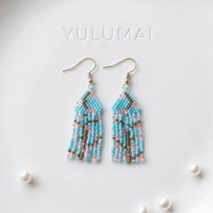 May include: Handmade beaded earrings with a turquoise, pink, and brown color scheme. The earrings feature a geometric design at the top and long fringe. The word "YULUMAI" is embossed on the white surface.