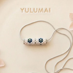 Peut inclure: Un collier de perles avec un pendentif cylindrique. Le pendentif présente un motif blanc, argenté et bleu. Le collier a une chaîne argentée et est présenté sur une surface crème. Le mot "YULUMAI" est estampé au-dessus du collier.