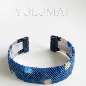 Peut inclure: Bracelet manchette bleu en perles avec intérieur en métal argenté. Le bracelet présente un motif floral avec des accents bleu clair et dorés. Le mot "YULUMAI" est affiché en arrière-plan.