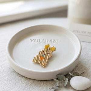 Peut inclure: Une broche lapin dorée en perles avec des oreilles jaunes et une carotte orange, posée sur un plat en céramique blanc. Le plat porte l'inscription "YULUMAI". Un accessoire décoratif.