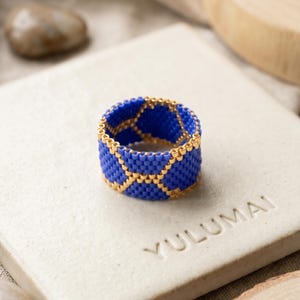 Peut inclure: Une bague artisanale avec un motif tissé de petites perles bleues et des accents dorés. La bague a une bande épaisse et un design géométrique. Le mot "YULUMAI" est visible sur la surface en dessous.