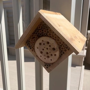 May include: A light-colored wooden bee house with a peaked roof, featuring a central circular log slice with drilled holes surrounded by numerous small bamboo tubes arranged within a square frame to provide nesting space for solitary bees.