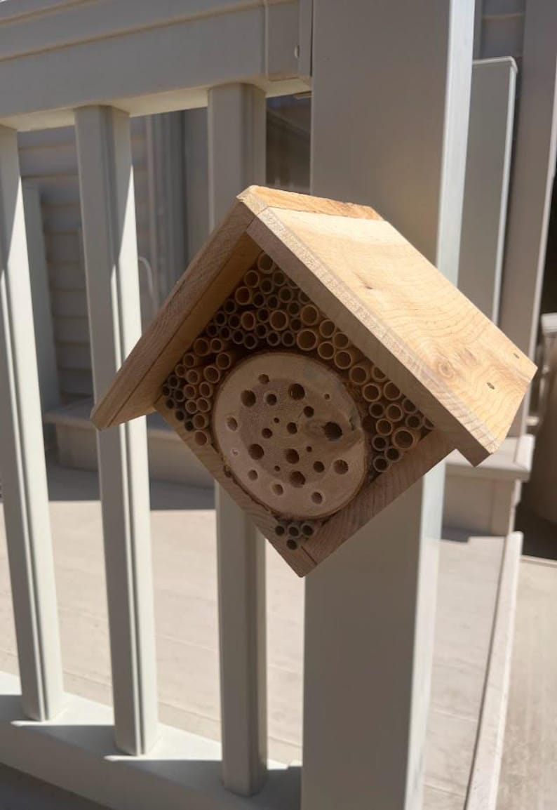 May include: A light-colored wooden bee house with a peaked roof, featuring a central circular log slice with drilled holes surrounded by numerous small bamboo tubes arranged within a square frame to provide nesting space for solitary bees.