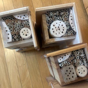 May include: Three wooden bee houses with open-front designs. Each structure contains a collection of hollow bamboo tubes and drilled wooden blocks with various circular openings, providing nesting cavities for solitary bees in a natural wood finish.