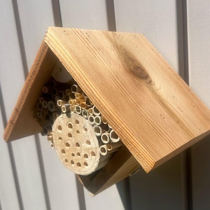 May include: A wooden bee house with a peaked roof, featuring a central circular wooden block with drilled holes surrounded by a collection of hollow bamboo tubes of varying diameters, all housed within a natural light brown cedar wood frame structure.