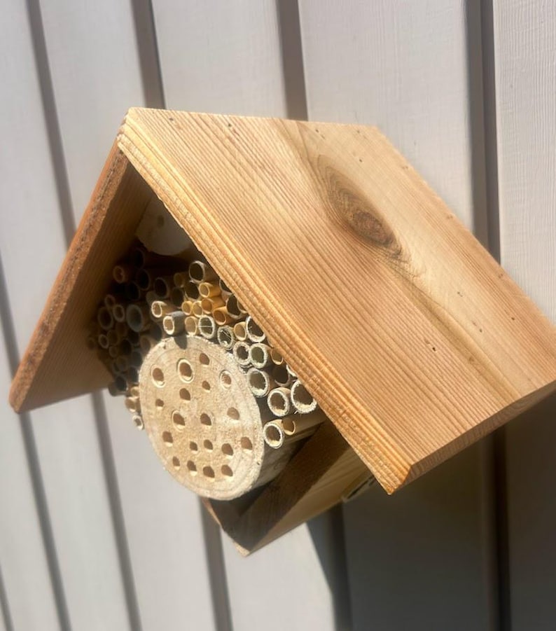 May include: A wooden bee house with a peaked roof, featuring a central circular wooden block with drilled holes surrounded by a collection of hollow bamboo tubes of varying diameters, all housed within a natural light brown cedar wood frame structure.