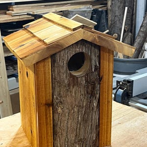 May include: A handcrafted wooden birdhouse with a natural bark front and a shingled roof. The birdhouse features a circular entrance hole and is constructed from various shades of brown wood.