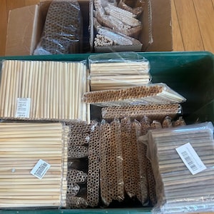 May include: A large green plastic bin and two cardboard boxes filled with numerous bundles of light-colored, cylindrical drinking straws wrapped in clear plastic. Some bundles are arranged horizontally, while others are stacked vertically.