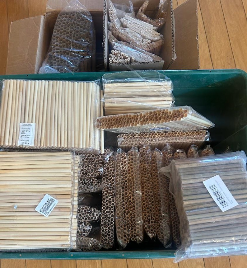 May include: A large green plastic bin and two cardboard boxes filled with numerous bundles of light-colored, cylindrical drinking straws wrapped in clear plastic. Some bundles are arranged horizontally, while others are stacked vertically.