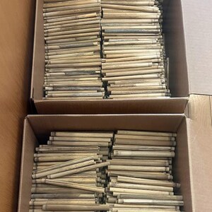 May include: Two cardboard boxes filled with a large quantity of light brown, cylindrical wooden sticks. Each stick features a natural wood grain texture and a smooth, straight finish, arranged in a dense, organized horizontal stack.