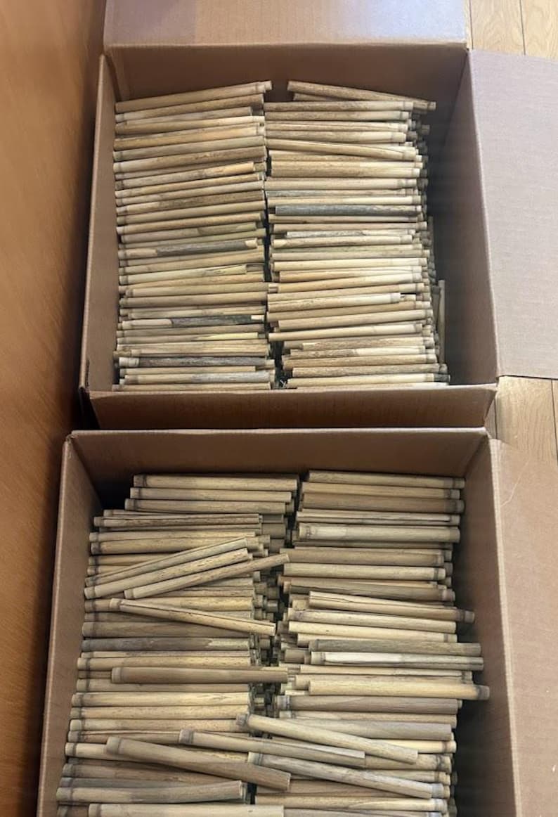 May include: Two cardboard boxes filled with a large quantity of light brown, cylindrical wooden sticks. Each stick features a natural wood grain texture and a smooth, straight finish, arranged in a dense, organized horizontal stack.