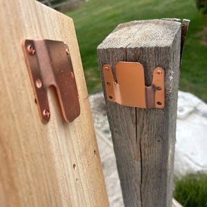 May include: Two copper-colored metal brackets are attached to wooden posts with screws. One bracket is on a light-colored, smooth wooden post, and the other is on a weathered, gray wooden post. The brackets have a U-shaped design.