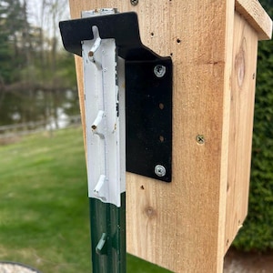 May include: A wooden birdhouse with a pitched roof is mounted on a green metal post. The birdhouse is made of light-colored wood and is attached to the post with a black metal bracket. The post has white and green paint.