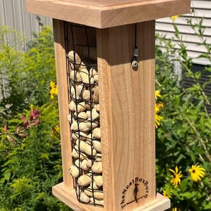 May include: A wooden bird feeder with a square top and base, and a rectangular body. The body features a wire mesh filled with peanuts. The feeder is suspended by black cords. The text "The Nest Box Barn" is visible on the side.