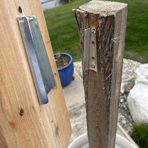 May include: Two wooden posts with metal brackets attached. One post is a light-colored, smooth plank, and the other is a rough-hewn, darker log. The brackets are silver and secured with screws, suggesting a construction or assembly project.