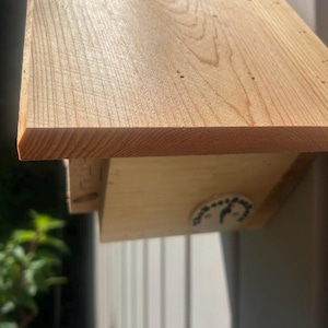 May include: A light-colored wooden birdhouse with a slanted roof and visible wood grain. A small circular white sticker with black text and a bird silhouette is attached to the side of the structure. The wood has a natural, unfinished appearance.