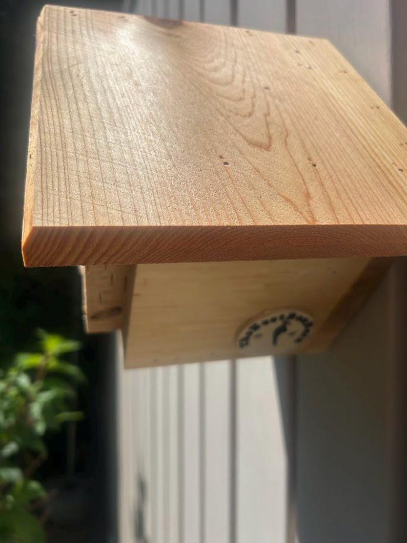 May include: A light-colored wooden birdhouse with a slanted roof and visible wood grain. A small circular white sticker with black text and a bird silhouette is attached to the side of the structure. The wood has a natural, unfinished appearance.
