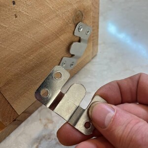 May include: A silver-colored metal Z-bar hanger bracket with three circular screw holes, held in front of a light brown wooden surface that features an identical metal bracket already attached with two visible screws.