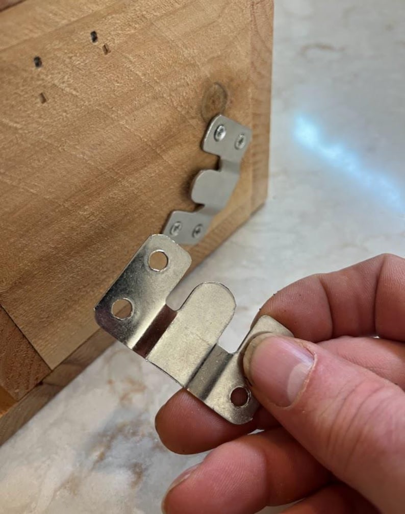 May include: A silver-colored metal Z-bar hanger bracket with three circular screw holes, held in front of a light brown wooden surface that features an identical metal bracket already attached with two visible screws.
