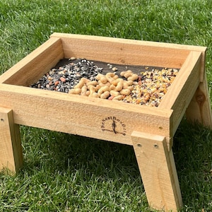 Puede incluir: Un comedero para pájaros de madera con una bandeja rectangular llena de semillas, cacahuetes y otros granos. El comedero tiene cuatro patas y está colocado sobre un césped. La marca "The Nest Box" es visible.