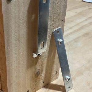May include: Two silver metal brackets, one attached to a wooden surface and the other lying on a wooden surface.