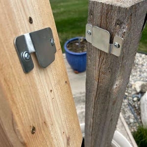 May include: Close-up of two wooden posts with metal brackets attached. One post is light-colored, the other is weathered gray. The brackets are silver and secured with screws, likely for joining or supporting structures. The background shows a lake and greenery.