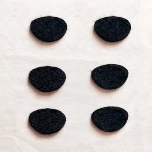 May include: Six small, black, oval-shaped felt pieces arranged in two rows of three on a light pink fabric background. The felt pieces have a slightly textured appearance.