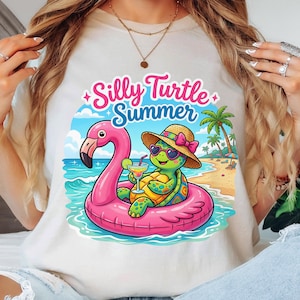 May include: A cream-colored t-shirt featuring a graphic of a green turtle wearing a straw hat and heart-shaped sunglasses, floating in a pink flamingo pool float. The turtle holds a cocktail. The text reads Silly Turtle Summer.