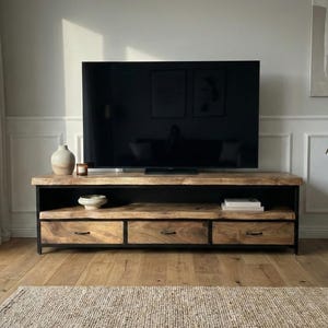 Puede incluir: Un mueble de televisi&oacute;n rectangular con una superficie de madera natural de borde vivo y tres cajones inferiores con herrajes de metal negro. La unidad est&aacute; sostenida por un marco de metal negro e incluye un estante abierto bajo la superficie.