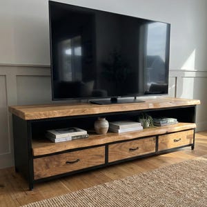 Puede incluir: Un mueble de televisi&oacute;n rectangular con una superficie y estante de madera natural, sostenido por un marco de metal negro. La unidad incluye tres cajones con tiradores de metal negro, proporcionando espacio de almacenamiento bajo el estante abierto.
