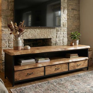 Puede incluir: Un mueble de televisi&oacute;n rectangular con una superficie de madera natural de borde vivo y tres cajones con tiradores de metal oscuro. La unidad cuenta con un marco de metal negro, un estante abierto para libros y una base de madera maciza.