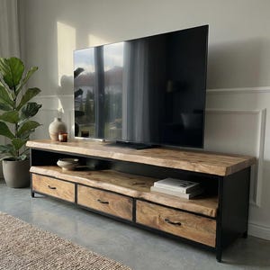Puede incluir: Un mueble de televisi&oacute;n rectangular largo con una superficie de madera natural de borde vivo y tres cajones con marcos y tiradores de metal negro. La unidad incluye un estante abierto para almacenamiento y se apoya sobre una base de metal negro.