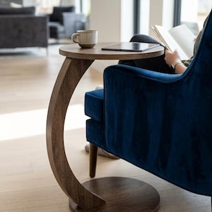 Könnte beinhalten: Ein Beistelltisch aus dunklem Holz mit runder Tischplatte und geschwungenem Bein. Eine kleine Tasse und ein Tablet liegen auf dem Tisch. Der Tisch steht neben einem blauen Samtsessel.