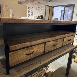 Puede incluir: Un mueble multimedia rectangular con marco de metal negro y tres cajones de madera con tiradores negros. La unidad incluye una superficie superior con borde natural y un estante de almacenamiento abierto situado bajo el tablero.