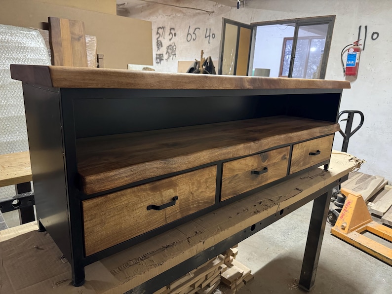 Puede incluir: Un mueble multimedia rectangular con marco de metal negro y tres cajones de madera con tiradores negros. La unidad incluye una superficie superior con borde natural y un estante de almacenamiento abierto situado bajo el tablero.