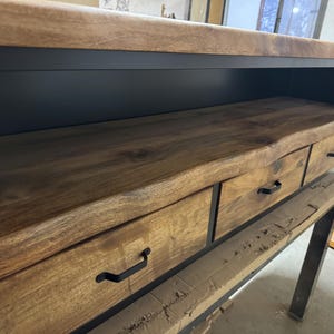 Puede incluir: Una mesa consola con una superficie de madera natural de borde vivo y tres cajones con tiradores de metal negro. La unidad est&aacute; sostenida por un marco de metal negro, mostrando una combinaci&oacute;n de texturas de grano de madera c&aacute;lida y estructura met&aacute;lica.