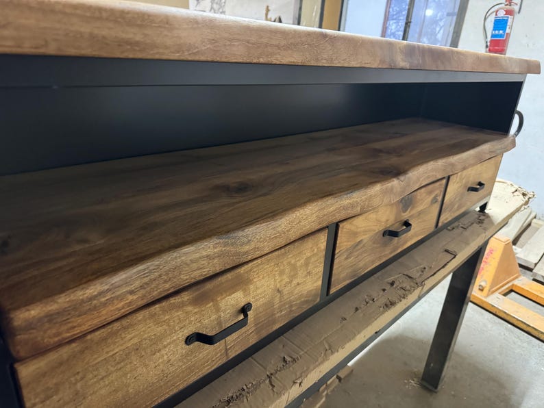 Puede incluir: Una mesa consola con una superficie de madera natural de borde vivo y tres cajones con tiradores de metal negro. La unidad est&aacute; sostenida por un marco de metal negro, mostrando una combinaci&oacute;n de texturas de grano de madera c&aacute;lida y estructura met&aacute;lica.