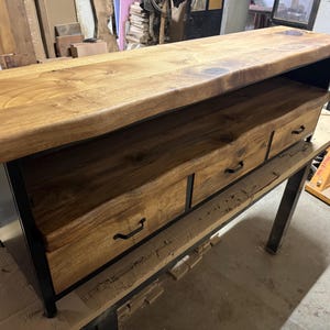 Puede incluir: Una mesa consola rectangular de madera con una tapa gruesa de borde natural y dos cajones con tiradores de metal negro. La unidad est&aacute; sostenida por un marco de metal negro, combinando un acabado de madera clara con una estructura industrial oscura.