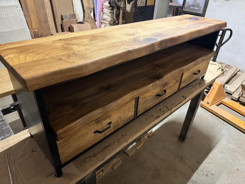 Puede incluir: Una mesa consola rectangular de madera con una tapa gruesa de borde natural y dos cajones con tiradores de metal negro. La unidad est&aacute; sostenida por un marco de metal negro, combinando un acabado de madera clara con una estructura industrial oscura.