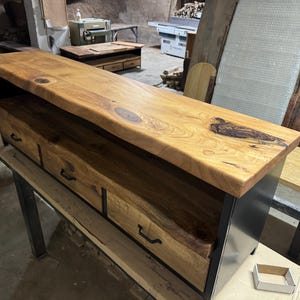Puede incluir: Un mueble multimedia rectangular de madera con una superficie de borde natural que muestra vetas y nudos visibles. La unidad incluye tres cajones frontales con tiradores de metal negro, integrados en una estructura de marco de metal negro.