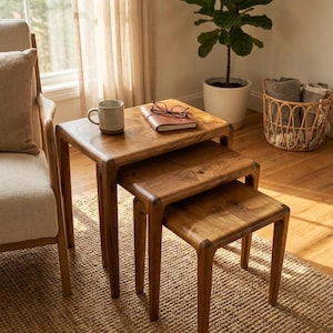 Könnte beinhalten: Dreiteiliges Set aus Holz-Nesting-Tischen mit natürlicher Oberfläche. Die Tische sind gestapelt angeordnet, um ihre unterschiedlichen Größen zu zeigen. Eine Tasse, eine Brille und ein Notizbuch liegen auf dem obersten Tisch. Die Szene spielt in einem hellen Wohnzimmer.