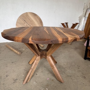 May include: A handcrafted wooden table with a unique, irregular edge. The tabletop is a mosaic of warm brown and tan wood grains. The table stands on a geometric, star-shaped base. The table's diameter is approximately 40 inches.