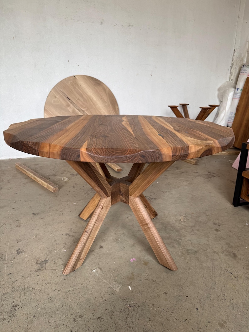 May include: A handcrafted wooden table with a unique, irregular edge. The tabletop is a mosaic of warm brown and tan wood grains. The table stands on a geometric, star-shaped base. The table's diameter is approximately 40 inches.